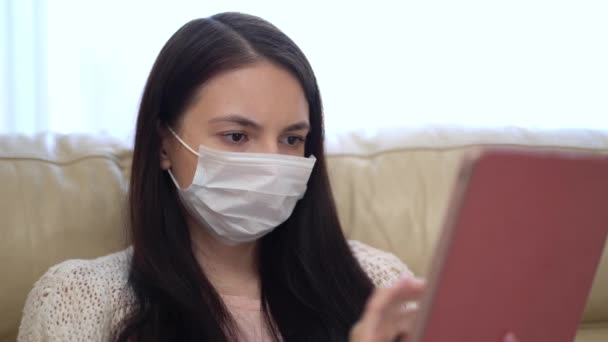 Kvinna i en medicinsk mask använder en tablett hemma under karantän. — Stockvideo