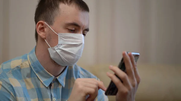 Man in a Medical Mask utiliza un teléfono inteligente en casa durante la cuarentena. Funciona solo desde casa de forma remota. Quarantine Covid-19 Corona Virus. Concepto de distanciamiento social . —  Fotos de Stock