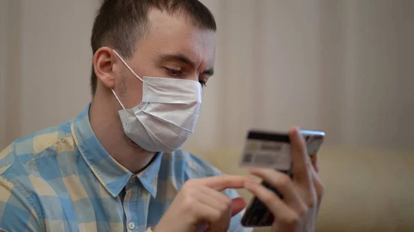 Cuarentena Covid-19 Coronavirus. Hombre con máscara de compras en línea con tarjeta de crédito utilizando el teléfono inteligente en casa. Entrega de alimentos, orden de pago. Distanciamiento social . —  Fotos de Stock