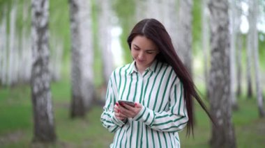 Elinde akıllı telefonuyla güzel bir kız baharda huş ağacı korusunda yürüyor.