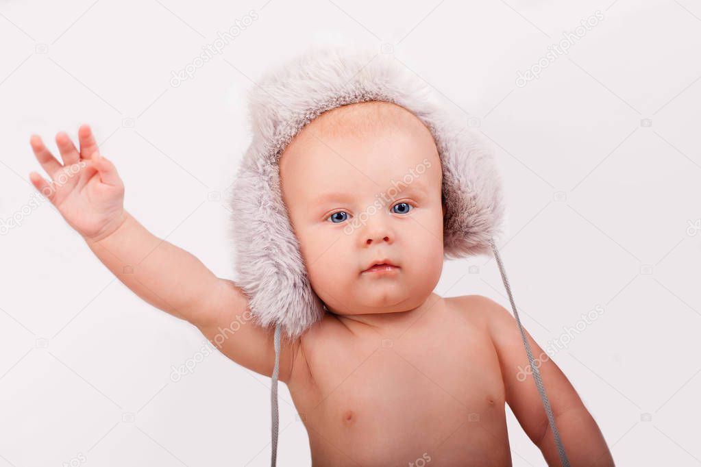 Portrait of cute caucasian baby nine month year old with blue eyes wearing a furry hat on a light background holding hand
