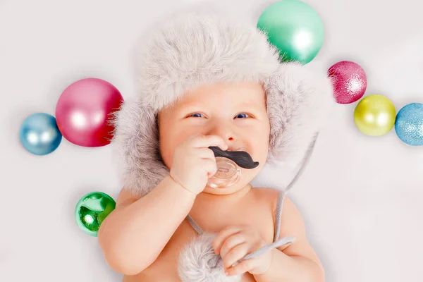 Niño lindo con un chupete en la forma de un bigote sosteniendo en su mano se encuentra en su espalda junto a los juguetes de Navidad. Sonriendo. . —  Fotos de Stock