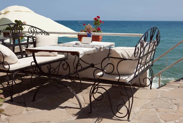 Nice outdoor cafe with forged furniture, white pillows and an awning at sunny summer day in resort town Sozopol, Bulgaria. Photo with natural sea background