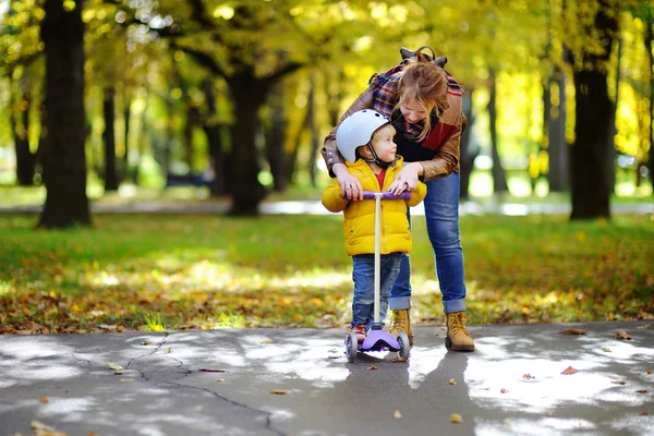 Junge Mutter zeigt ihrem kleinen Sohn, wie man einen Roller in einem herbstlichen Park fährt — Stockfoto