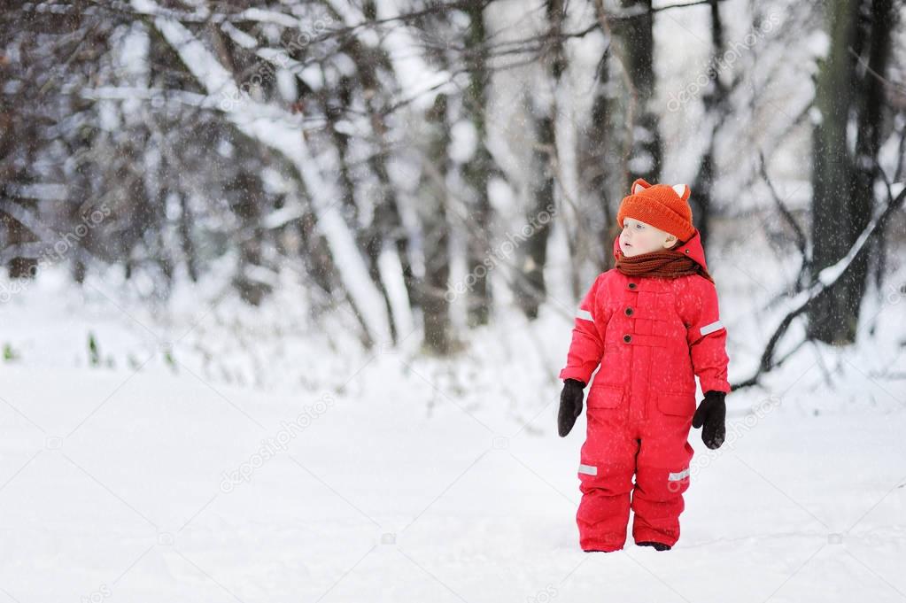 Portrait of little funny boy in red winter clothes having fun with snow during snowfall