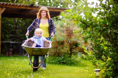 Adorable toddler boy having fun in a wheelbarrow pushing by mum in domestic garden clipart
