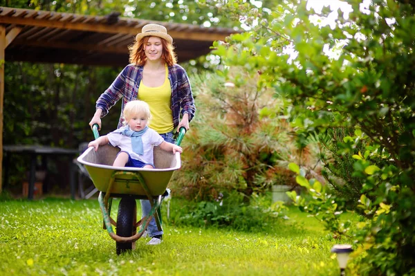 Чарівний маленький хлопчик розважається в інвалідному візку штовхаючи маму в домашньому саду — стокове фото