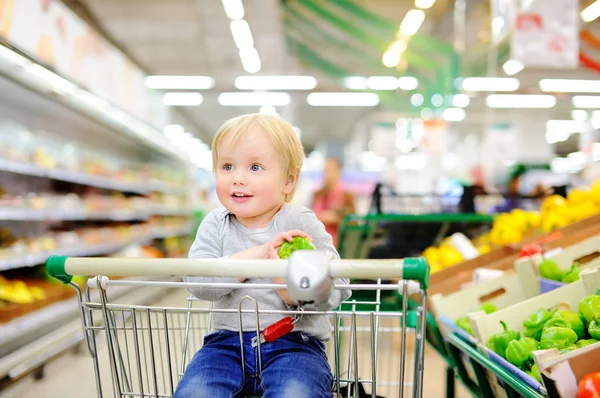 Söt liten knatte pojke sitter i kundvagnen i en stormarknad — Stockfoto