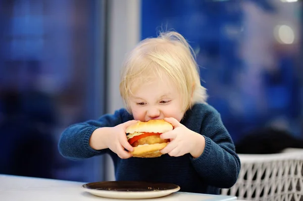 Bonito menino loiro comer hambúrguer no restaurante fast food — Fotografia de Stock