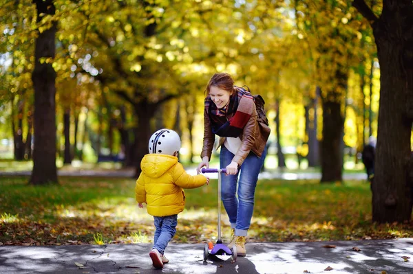 どのように秋の公園でスクーターに乗る彼女の幼児の息子を示す若い母親 — ストック写真