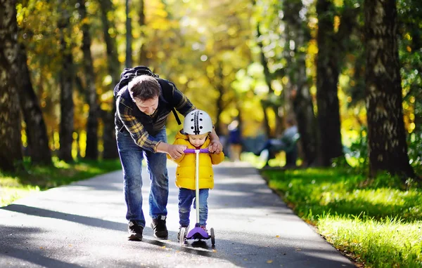 Middle age father showing his toddler son how to ride a scooter in a autumn park — Stock Photo, Image
