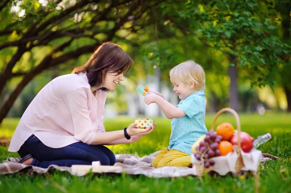 Cute little boy with his young mother opening nicely wrapped gift during picnic in sunny park — Stock Photo, Image