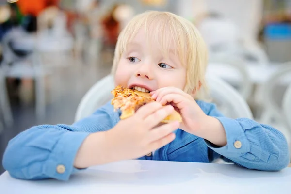 Lindo chico rubio comiendo rebanada de pizza en restaurante de comida rápida —  Fotos de Stock
