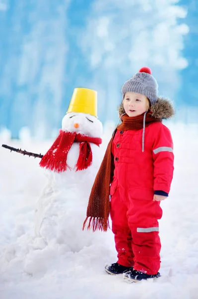 Niño con ropa roja de invierno divirtiéndose con muñeco de nieve en el parque nevado —  Fotos de Stock