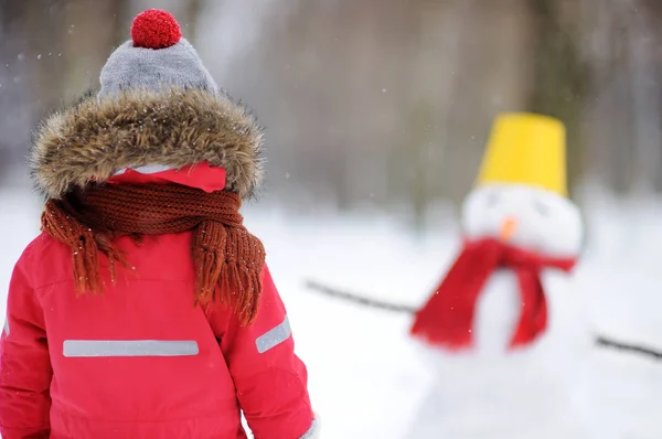 Anak kecil dengan pakaian musim dingin merah bersenang-senang dengan manusia salju di taman bersalju — Stok Foto