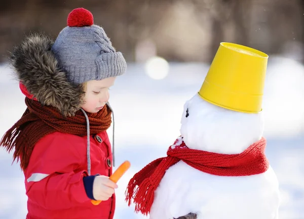 Menino em roupas vermelhas de inverno se divertindo com boneco de neve no parque nevado — Fotografia de Stock