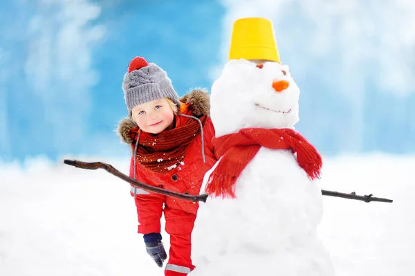 Μικρό αγόρι στο κόκκινο χειμωνιάτικα ρούχα διασκεδάζοντας με τον χιονάνθρωπο στο χιονισμένο πάρκο — Φωτογραφία Αρχείου