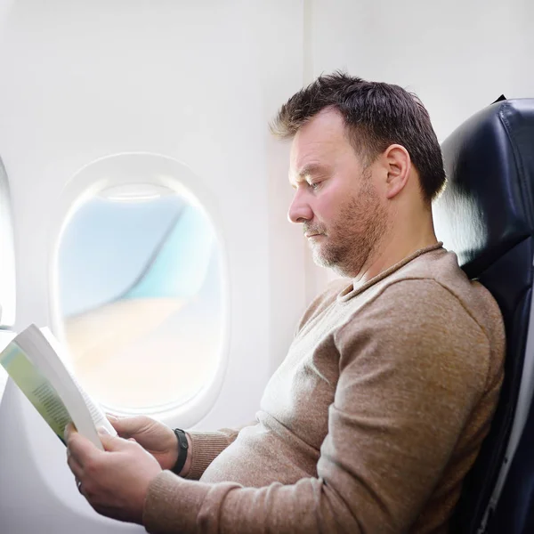 Мужчина средних лет путешествует на самолете и читает книгу во время полета. Транспортная концепция — стоковое фото