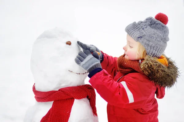 Menino em roupas vermelhas de inverno se divertindo com boneco de neve no parque nevado — Fotografia de Stock