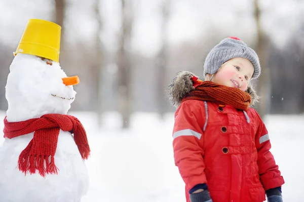 Menino em roupas vermelhas de inverno se divertindo com boneco de neve no parque nevado — Fotografia de Stock