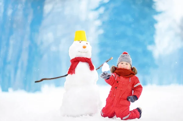 Menino em roupas vermelhas de inverno se divertindo com boneco de neve no parque nevado — Fotografia de Stock