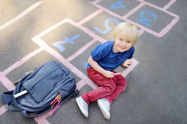 Cute blond boy playing hopscotch game after school with bags laying near — Stock Photo, Image