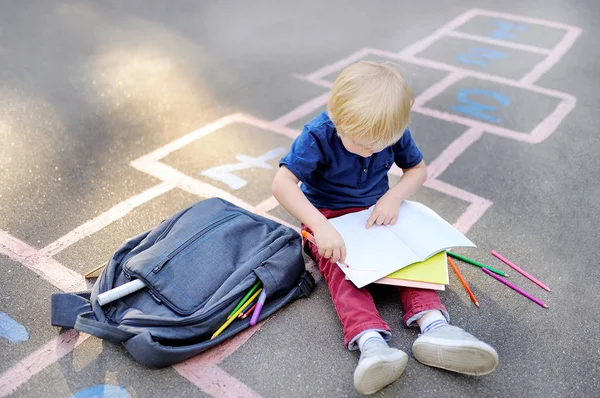 Милый блондин делает домашнее задание сидя на школьном дворе после школы с сумками, лежащими рядом — стоковое фото
