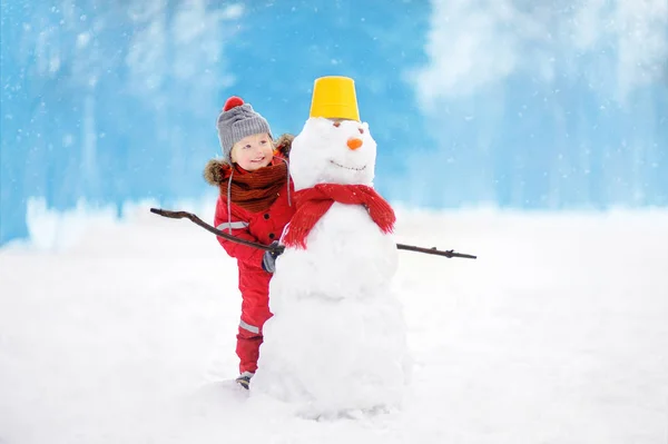 Niño con ropa roja de invierno divirtiéndose con muñeco de nieve en el parque nevado —  Fotos de Stock