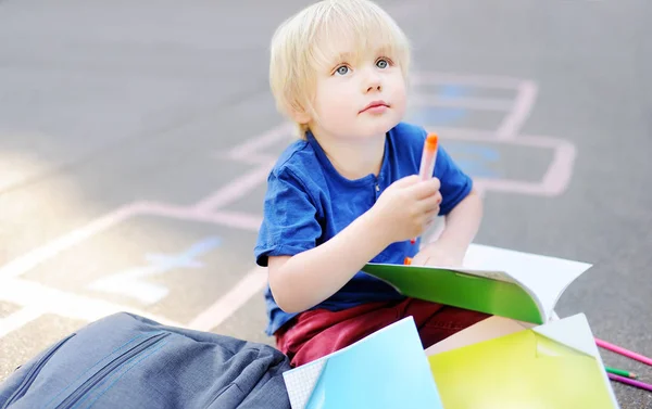 Mignon blond garçon faire des devoirs assis sur cour de l'école après l'école avec sacs couché près . — Photo