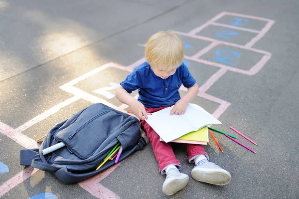 Милый блондин делает домашнее задание сидя на школьном дворе после школы с сумками, лежащими рядом . — стоковое фото