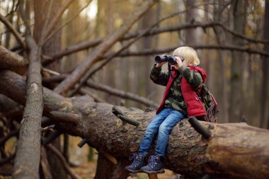 Sonbahar ormanlarında yürüyüş yaparken dürbünlü küçük bir izci. Çocuk büyük bir ağacın üzerinde oturuyor ve dürbünle bakıyor..