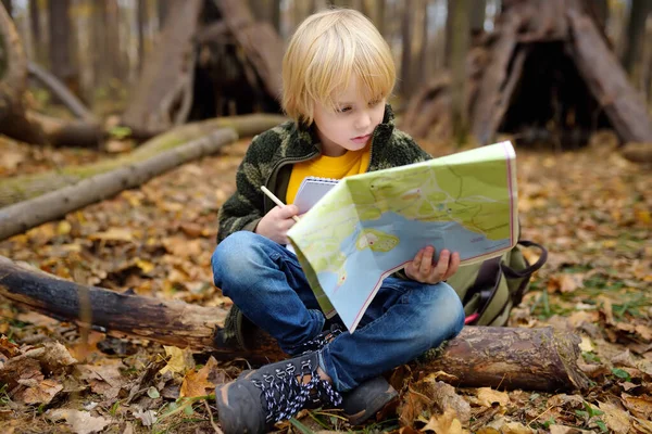 Malý skaut se orientuje v lese. Dítě sedí na spadlém stromě a dívá se na mapu na pozadí teepee chaty. — Stock fotografie