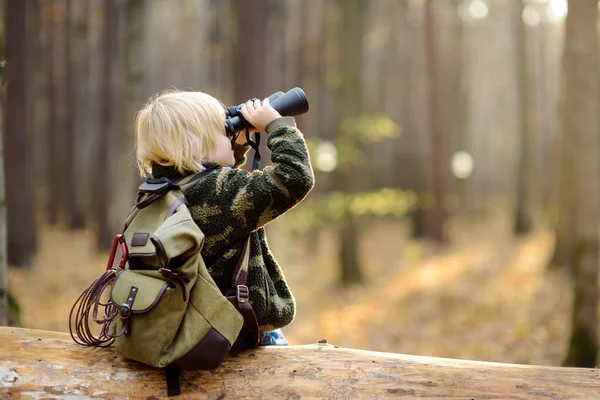 Niño explorador con binoculares durante el senderismo en el bosque de otoño. Niño está sentado en un gran árbol caído y mirando a través de un prismáticos . —  Fotos de Stock