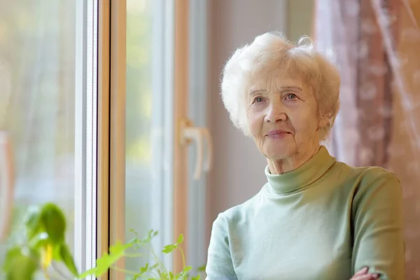 Porträt einer schönen lächelnden Seniorin mit lockigem weißem Haar. ältere Dame steht zu Hause am Fenster. — Stockfoto