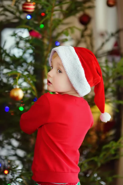 小男孩戴着圣诞礼帽,准备庆祝圣诞节. 可爱的孩子用玻璃玩具装饰圣诞树 — 图库照片
