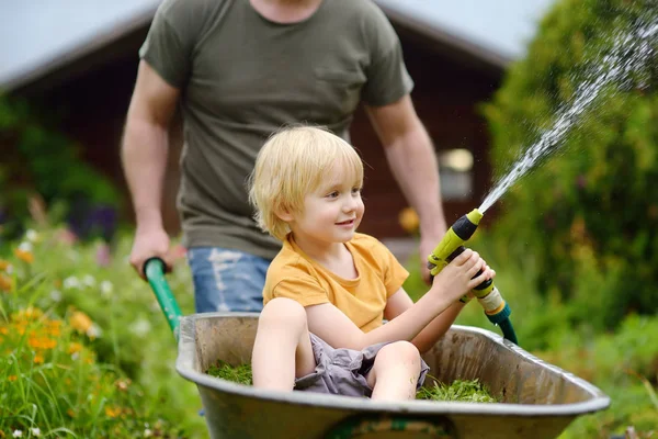 Niño feliz divirtiéndose en una carretilla empujando por papá en el jardín doméstico en el día soleado cálido. Plantas de riego para niños de una manguera . — Foto de Stock