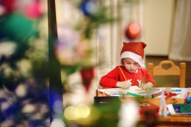 Noel Baba kostümü giymiş küçük bir çocuk süslü bir Noel ağacının yanında çizim yapıyor..