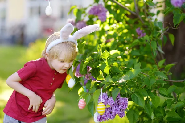 Красивый маленький мальчик в кроличьих ушах охотится за пасхальными яйцами в весеннем парке на Пасху . — стоковое фото