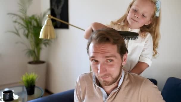 若いハンサムな父親のための面白い髪型を作る小さな巻き毛の娘 未就学児の女の子は父親の髪を磨き 美容室で働いているかのように遊んでいます 幸せな親 — ストック動画
