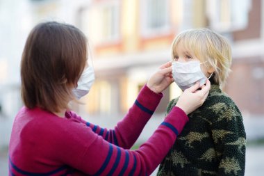 Koruyucu maske takan genç bir kadın havaalanında, süpermarkette ya da başka bir yerde oğluna maske takıyor. COVID-19 salgını sırasında güvenlik. Virüs salgını..