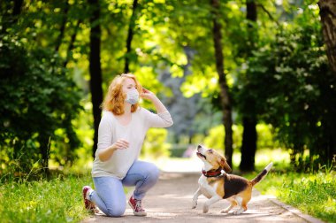 Coronavirus salgını sırasında parkta Beagle köpeğiyle oynayan tek kullanımlık tıbbi maske takan güzel bir kadın. Evcil hayvan yürüyüşü. Covid-19 salgını devam ederken kamusal alanda güvenlik.