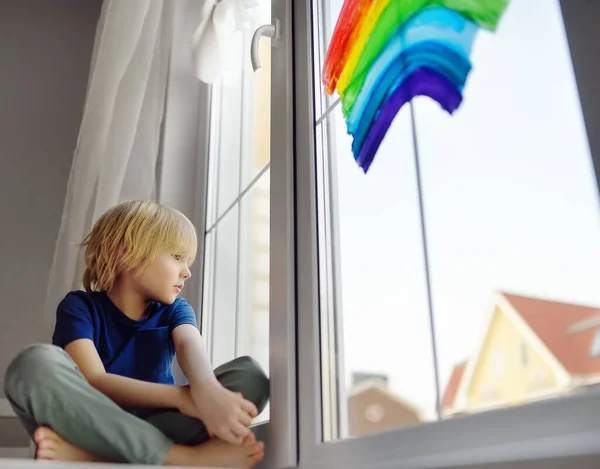コロナウイルスの隔離中に虹を描くと窓に座っている小さな男の子 レインボーサインは希望の象徴ですすべてがうまくいくことを意味しますコロナウイルスをロックダウンするために家にいてください — ストック写真
