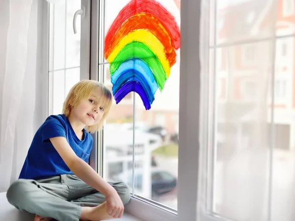 Little boy sitting at window with drawing rainbow while coronavirus quarantine. Rainbow sign is symbol of hope, means that everything will be OK. Stay at home for lockdown coronavirus.