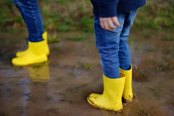 Mischievous preschooler child and adult wearing yellow rubber rain boots jumping in large wet mud puddle. Person playing and having fun. Outdoors games in spring.