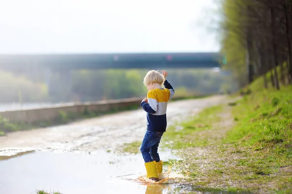 雨の後に大きなぬれた泥の水たまりにジャンプする黄色のゴム製の雨のブーツを身に着けているいたずらっ子 晴れた春の日に子供が遊んで楽しんでいます 子供のための屋外ゲーム — ストック写真