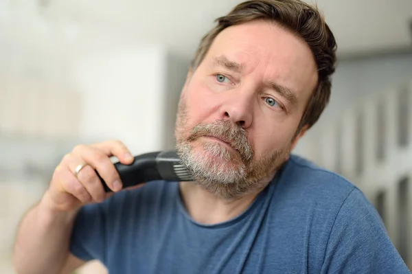 在检疫期间 一个成熟的男人正在家里用电动剃须刀刮胡子 理发店关门时 英俊的大胡子男人在家里用修整器修剪胡子 — 图库照片