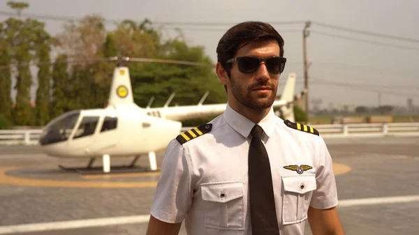 Portrait of commercial pilot in uniform standing near small private helicopter