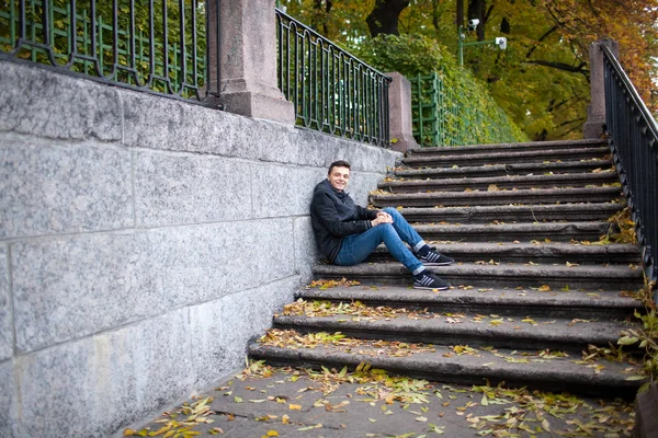 Ein junger Kerl sitzt auf der Treppe im Park zwischen den gelben Blättern — Stockfoto