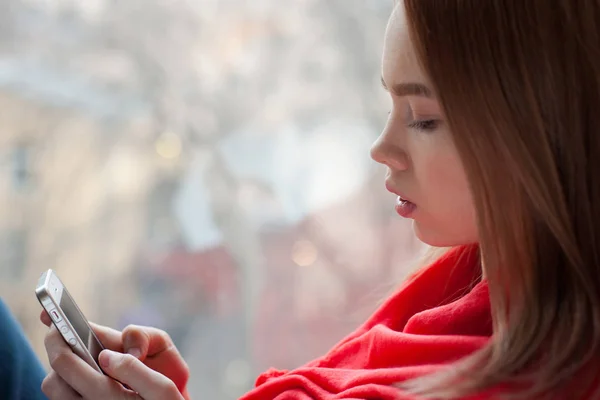 Retrato de uma menina lendo uma mensagem em um telefone sentado à janela — Fotografia de Stock