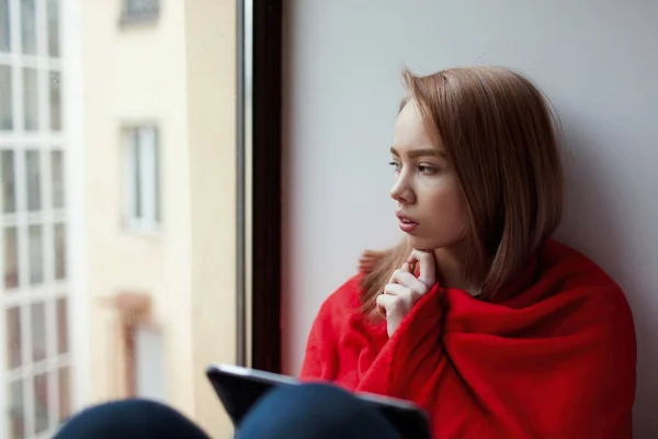 Ein junges Mädchen sitzt mit einem E-Book am Fenster, eingehüllt in eine rote Decke und denkt — Stockfoto